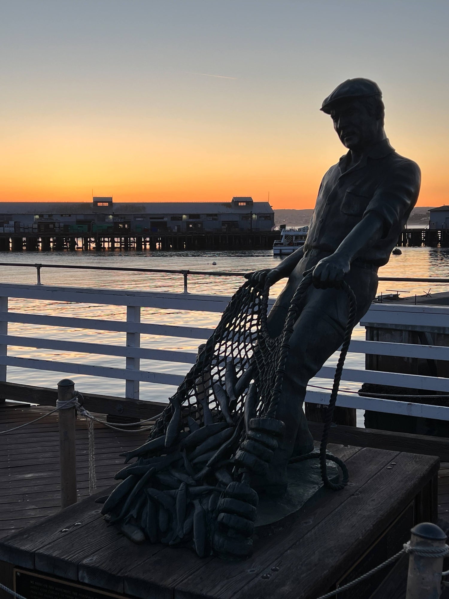 A fisherman's statue at the wharf in Monterey at sunrise with another pier in the background.